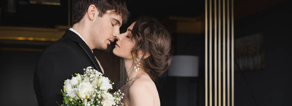 Seitenansicht des gut aussehenden Bräutigams in schwarz formale tragen küssende brünette Braut mit luxuriösem Schmuck hält Brautstrauß von Blumen, während sie in der Hotellobby stehen, Banner  - Foto, Bild