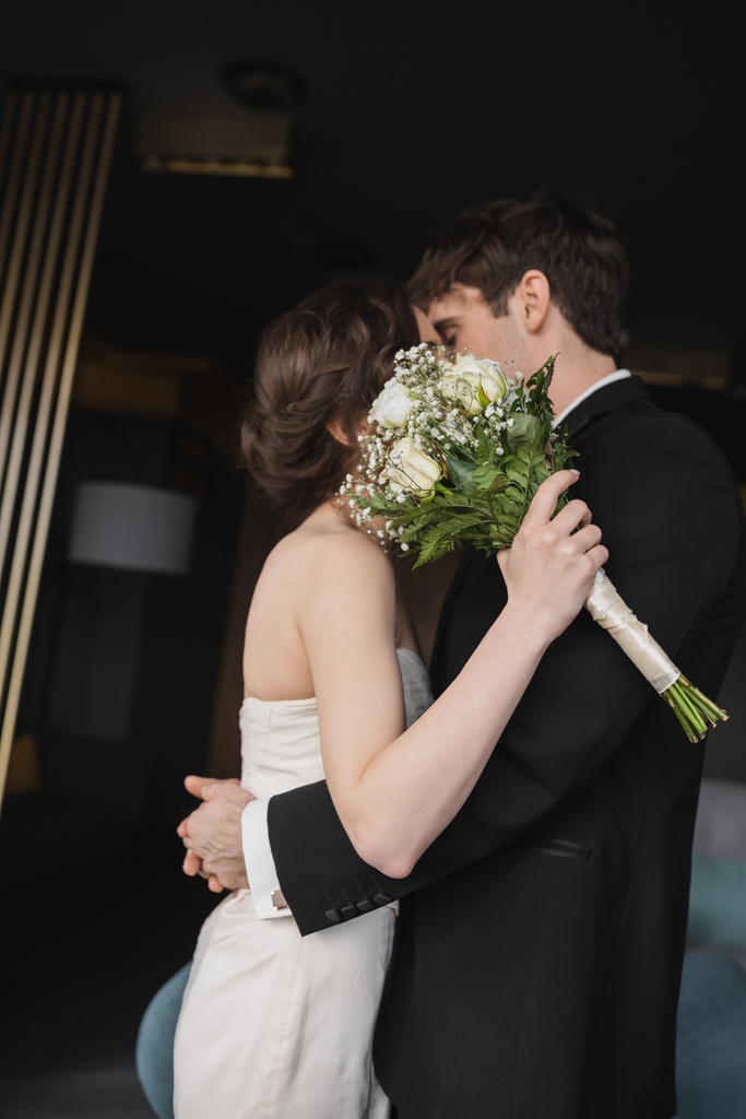 брюнетка наречена в елегантній весільній сукні тримає весільний букет з квітами і обличчями, цілуючись з нареченим в чорному формальному одязі, стоячи в готельному лобі
  - Фото, зображення
