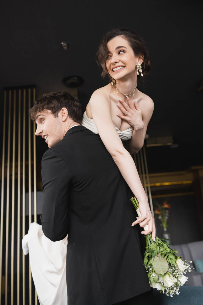 dobře vypadající ženich v černé formální oblečení zvedání veselá nevěsta s make-upem v bílých svatebních šatech drží svatební kytice květin, zatímco stojí v hotelové hale  - Fotografie, Obrázek