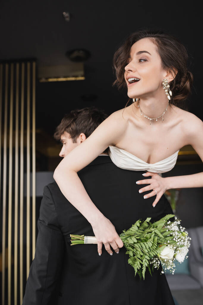 νεαρός γαμπρός σε μαύρο επίσημο ένδυμα άρση χαρούμενη νύφη σε λευκό νυφικό με ανοιχτό το στόμα κρατώντας νυφικό μπουκέτο λουλούδια, ενώ στέκεται στο λόμπι του ξενοδοχείου  - Φωτογραφία, εικόνα