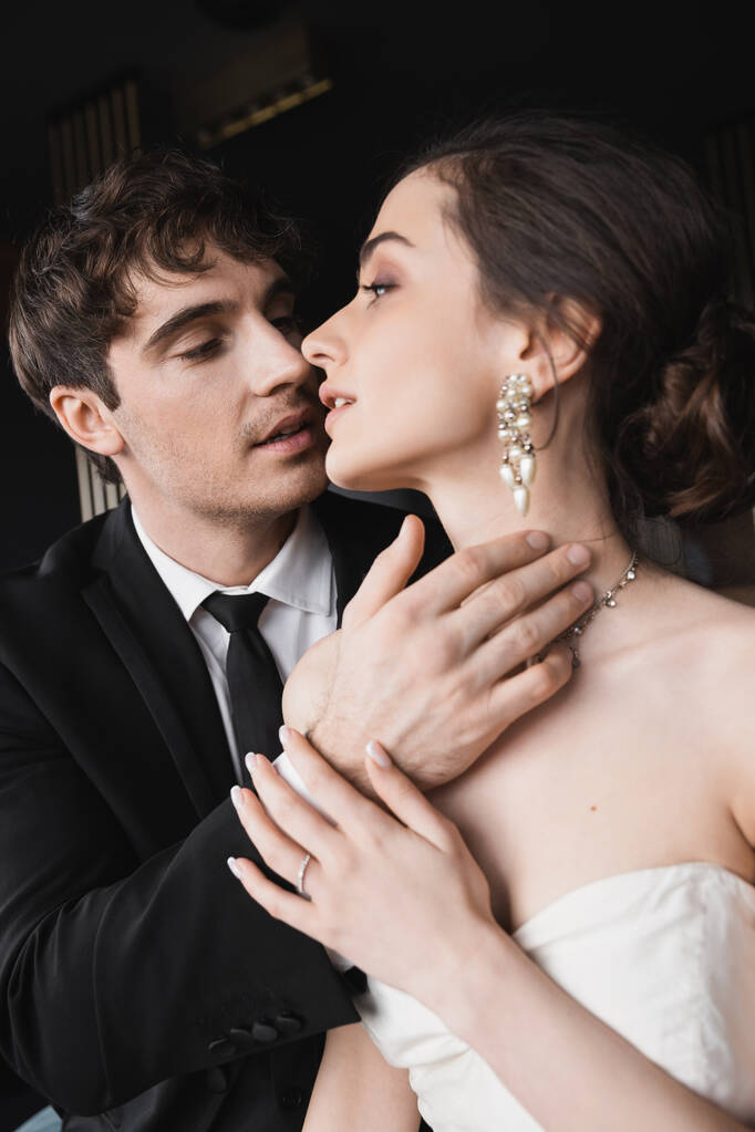 portrait de marié tendre en costume noir avec cravate toucher le visage de charmante mariée en robe de mariée blanche et bijoux tout en se regardant dans la chambre d'hôtel  - Photo, image