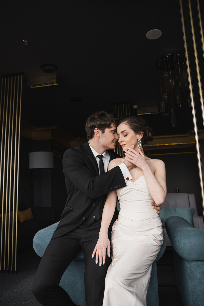 glücklicher Bräutigam im schwarzen Anzug mit Krawatte berührt Gesicht der charmanten Braut in weißem Brautkleid und Schmuck auf blauem Sofa in modernem Hotelzimmer gelehnt  - Foto, Bild