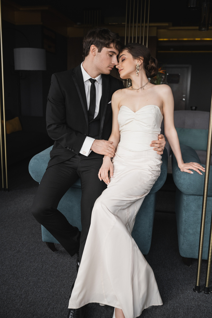τρυφερός γαμπρός σε μαύρο κοστούμι αγκαλιάζει τη μέση της γοητευτικής νύφης σε λευκό νυφικό και κοσμήματα, ενώ κλίνει μαζί σε μπλε καναπέ στο σύγχρονο δωμάτιο ξενοδοχείου  - Φωτογραφία, εικόνα