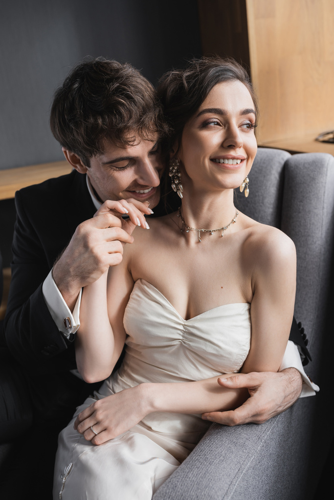 vrolijke bruidegom in zwart pak hand in hand van vrolijke bruid in witte jurk en luxe sieraden, terwijl glimlachen en zitten samen op comfortabele fauteuil in hotelkamer  - Foto, afbeelding