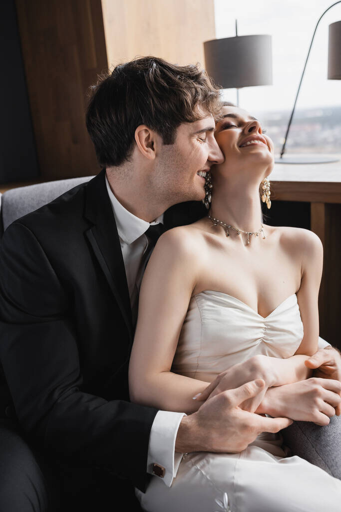 счастливый жених в черном костюме обнимает веселую невесту в белом платье и роскошные украшения, улыбаясь и сидя вместе на удобном кресле в гостиничном номере  - Фото, изображение