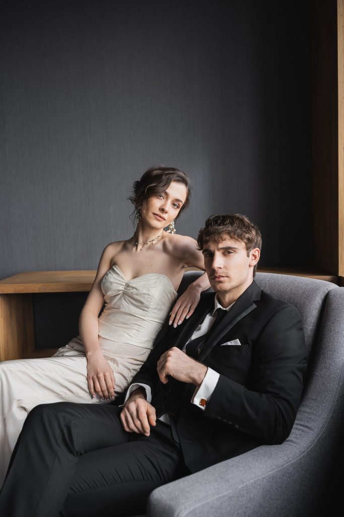 brünette Frau in weißem Hochzeitskleid und luxuriösem Schmuck sitzt auf bequemen Sesseln zusammen mit gut aussehendem Bräutigam im schwarzen Anzug im Hotelzimmer  - Foto, Bild