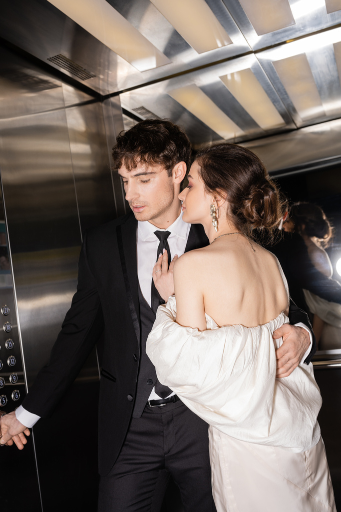 novio de buen aspecto en el botón de desgaste formal mientras abraza morena y novia joven en vestido de novia y de pie juntos en ascensor de hotel moderno  - Foto, imagen