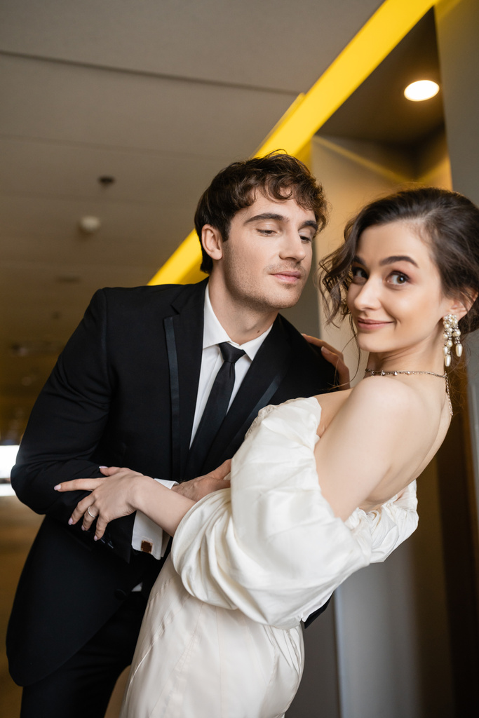 весёлый мужчина в черном костюме склоняется к великолепной невесте в белом свадебном платье, улыбаясь вместе и стоя в коридоре современного отеля, концепция медового месяца  - Фото, изображение