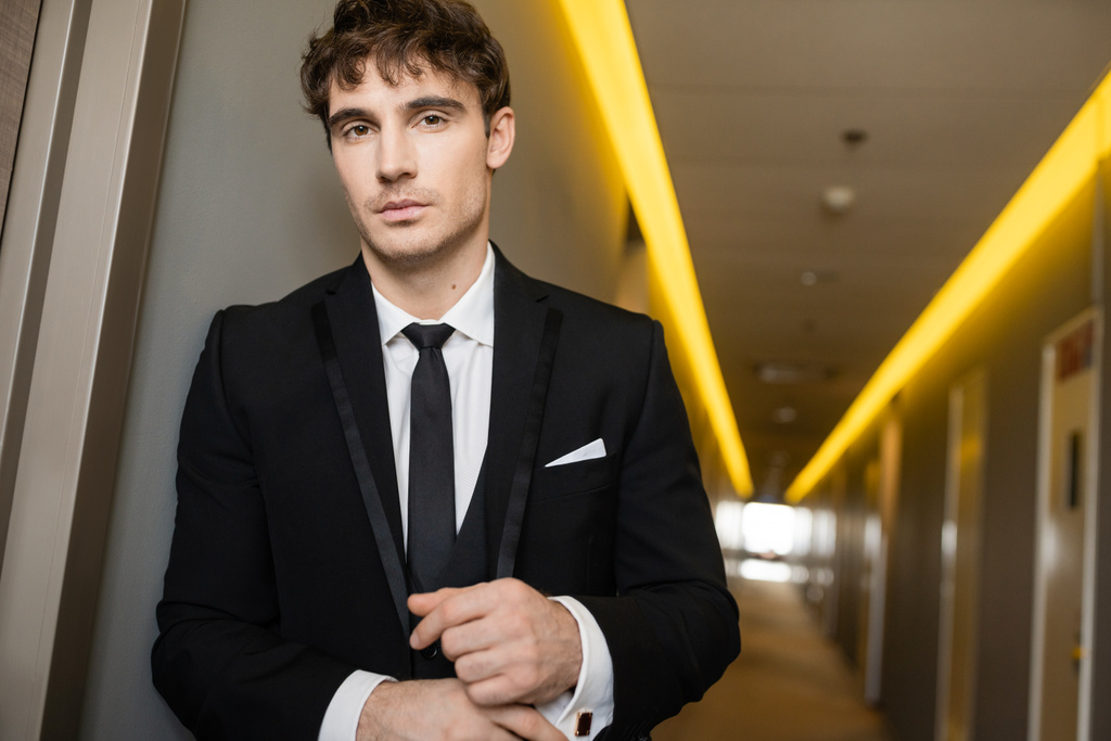 portrait of good looking young groom in black suit with while shirt and tie looking at camera while standing in corridor of modern hotel on wedding day  - Photo, Image