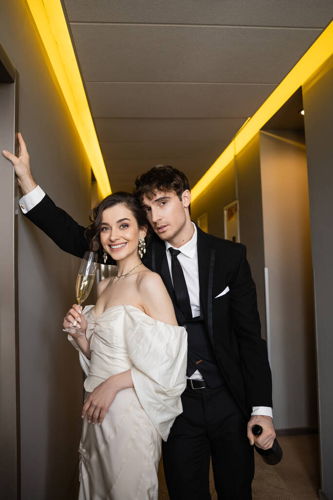 good looking groom in black formal wear leaning towards wall and holding bottle near happy bride with glass of champagne while standing together in hallway of modern hotel  - Photo, Image