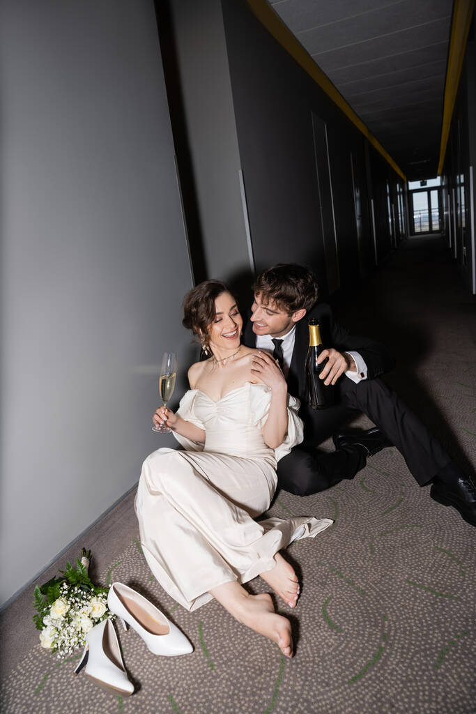 boldog vőlegény fekete öltönyben kezében üveg és ül közel gyönyörű menyasszony egy pohár pezsgővel mellett menyasszonyi csokor és magassarkú a padlón a folyosón a modern szálloda, friss házasok nászúton  - Fotó, kép