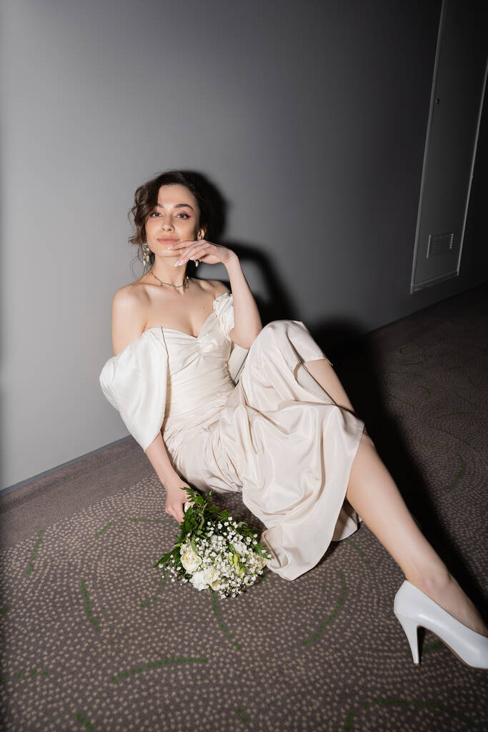 junge Braut mit brünetten Haaren im weißen Hochzeitskleid lächelnd und in die Kamera blickend, während sie neben einem Brautstrauß mit Blumen auf dem Boden der Halle in einem modernen Hotel sitzt  - Foto, Bild