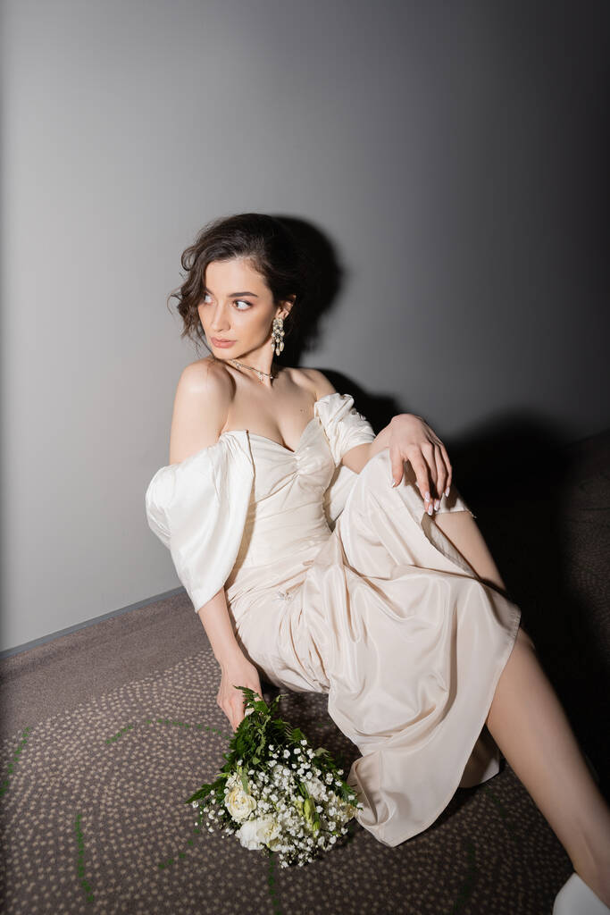 jovem noiva radiante com cabelo morena em vestido de noiva branco olhando para longe enquanto segurando buquê de noiva com flores e sentado no chão do salão no hotel moderno  - Foto, Imagem