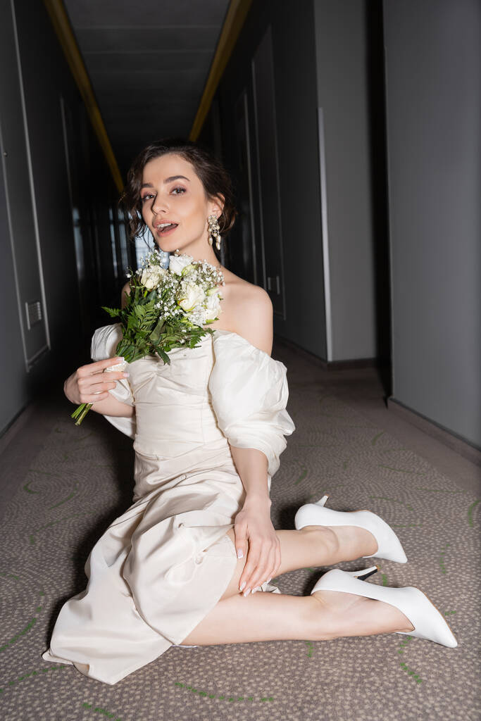amazed young bride with brunette hair in white wedding dress looking at camera while holding bridal bouquet with flowers and sitting on floor of hallway in modern hotel  - Photo, Image
