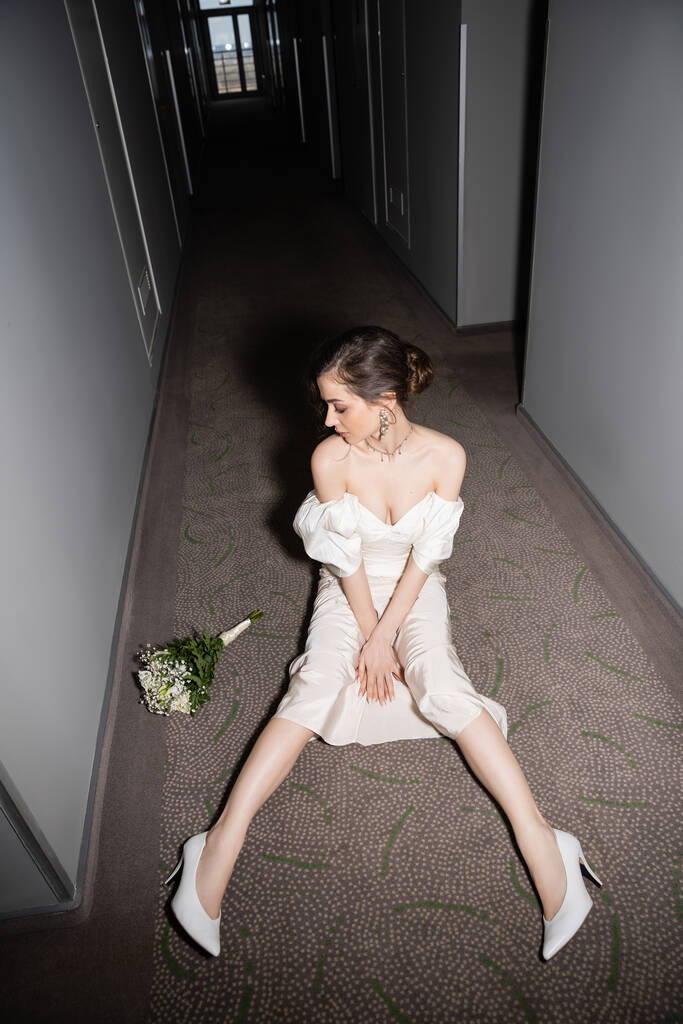 Beyaz gelinlik ve yüksek topuklu ayakkabılarla genç gelinin modern otelin koridorunda otururken elinde çiçeklerle gelin buketine baktığı yüksek açılı manzara.  - Fotoğraf, Görsel