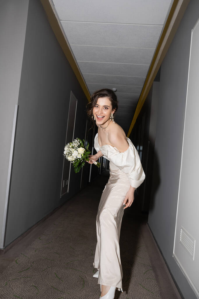 ενθουσιασμένοι και μελαχρινή νεαρή νύφη σε λευκό νυφικό χαμογελώντας, ενώ κρατώντας νυφικό μπουκέτο με λουλούδια και κοιτάζοντας κάμερα στην αίθουσα στο σύγχρονο ξενοδοχείο, όμορφη νύφη  - Φωτογραφία, εικόνα
