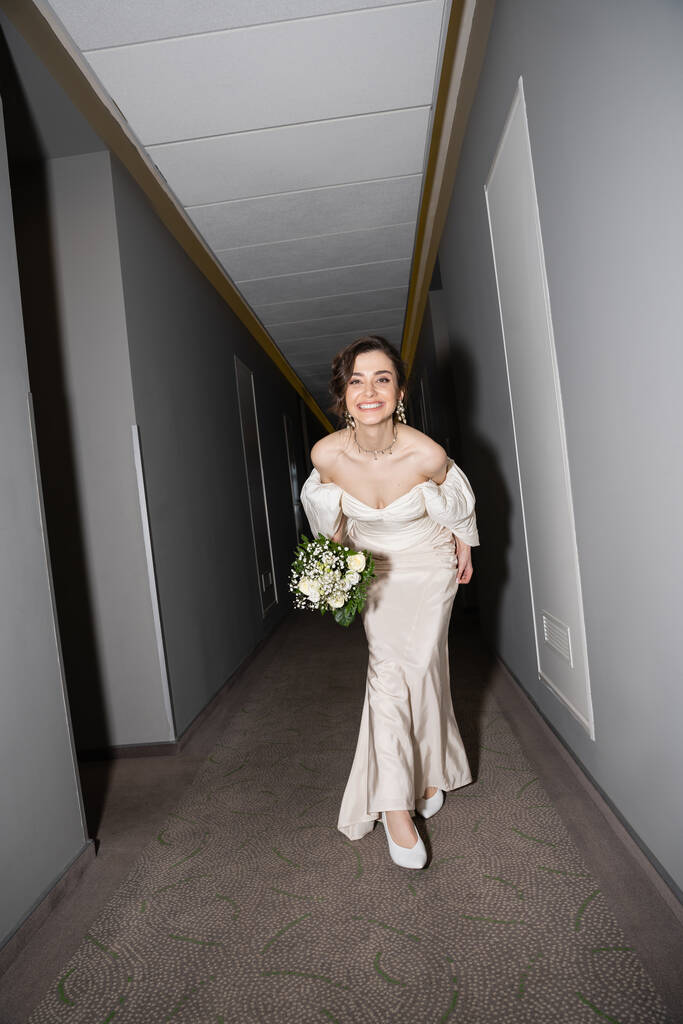 pleine longueur de jeune mariée excitée et brune en robe de mariée blanche souriante tout en tenant bouquet nuptiale avec des fleurs et en regardant la caméra dans le hall de l'hôtel moderne  - Photo, image