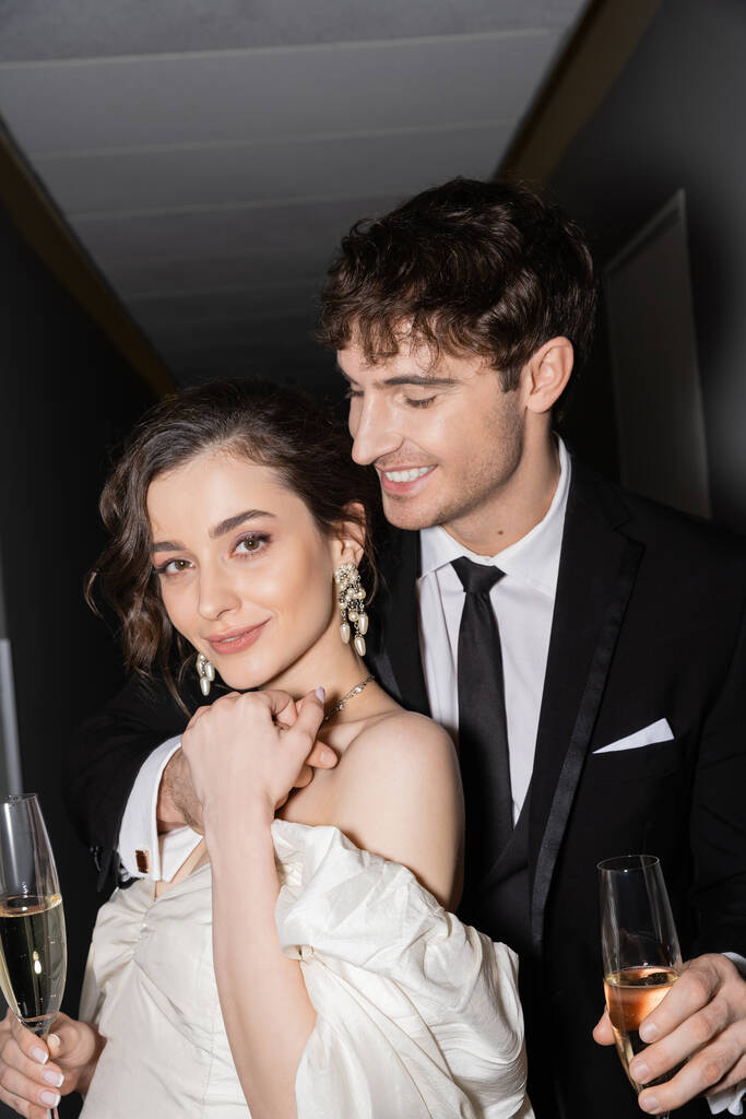 白のウェディングドレスを着た若いブルネットの花嫁と陽気な新郎がホテルの廊下で一緒に立って笑っている間にシャンパンのグラスを持っている  - 写真・画像