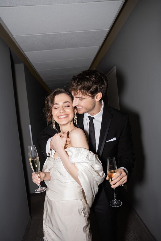 young and joyful bride in white wedding dress and cheerful groom holding glasses of champagne while standing and smiling together in hallway of hotel, newlyweds on honeymoon  - Photo, Image