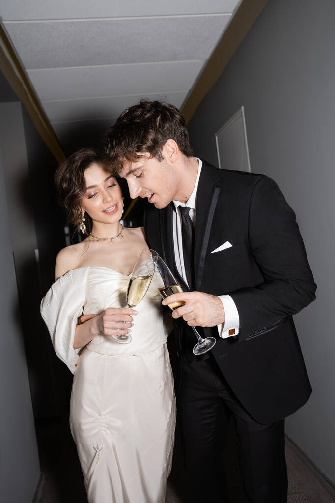 陽気な新郎とブルネットの花嫁白いウェディングドレスとシャンパンのクリンクグラスホテルの廊下で一緒に立って笑顔、新婚旅行で新婚旅行  - 写真・画像