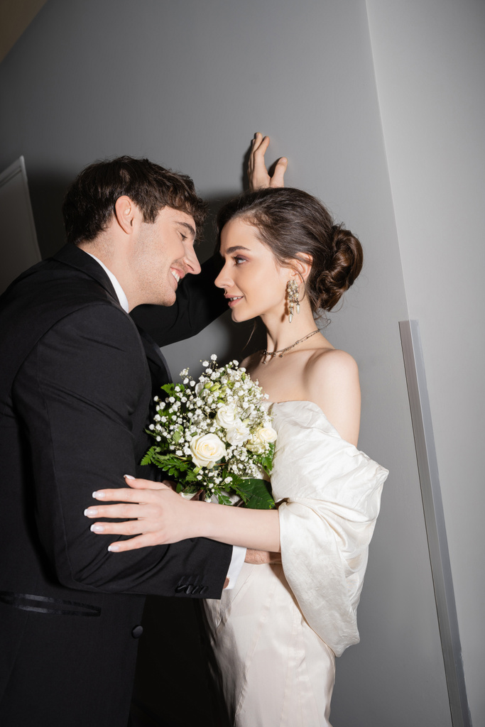 χαρούμενος γαμπρός σε μαύρο κοστούμι κλίνει προς τον τοίχο και κοιτάζοντας νύφη σε λευκό νυφικό κρατώντας νυφικό μπουκέτο, ενώ στέκεται μαζί στο διάδρομο του σύγχρονου ξενοδοχείου  - Φωτογραφία, εικόνα