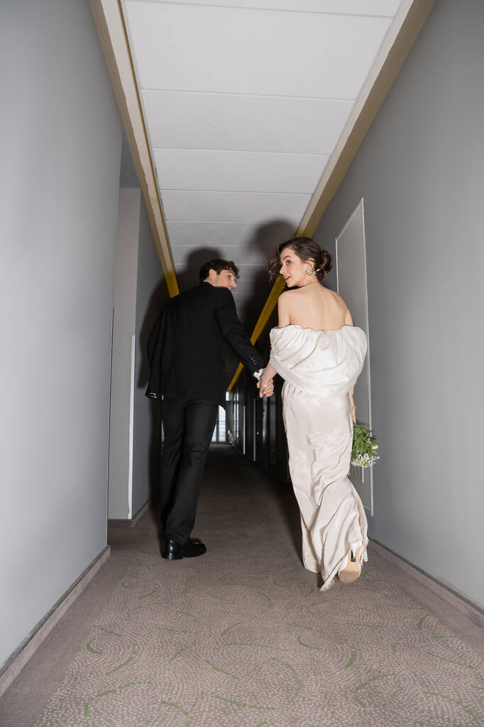 vista de ángulo bajo del novio en traje negro cogido de la mano con la novia en vestido de novia blanco llevando ramo de novia mientras caminan juntos en el pasillo del hotel moderno, recién casados en luna de miel  - Foto, imagen