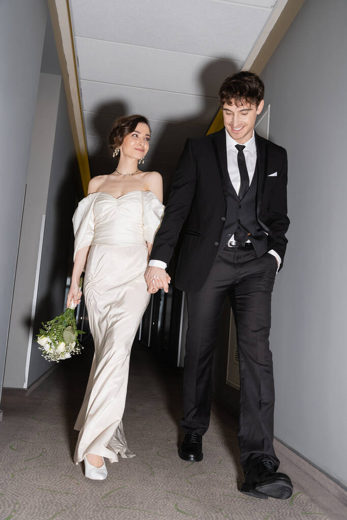 χαμηλή γωνία άποψη του χαρούμενου γαμπρού σε μαύρο κοστούμι ποζάρουν με το χέρι στην τσέπη και κρατώντας το χέρι της νύφης σε λευκό νυφικό που μεταφέρουν νυφικό μπουκέτο, ενώ το περπάτημα μαζί στην αίθουσα του σύγχρονου ξενοδοχείου  - Φωτογραφία, εικόνα