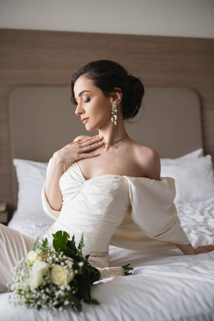 betoverende jonge bruid in witte jurk en luxe sieraden zitten op bed naast bruids boeket met bloemen in de moderne slaapkamer van het hotel kamer op trouwdag  - Foto, afbeelding