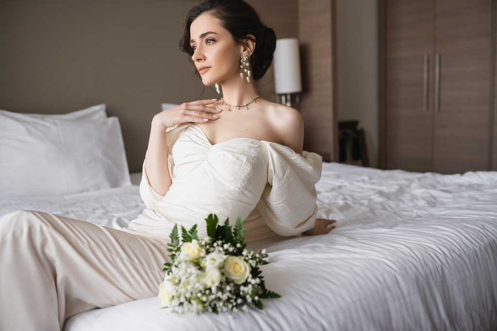 incantevole sposa in abito bianco e gioielli di lusso seduta sul letto accanto al bouquet da sposa e guardando lontano in camera da letto moderna in camera d'albergo il giorno del matrimonio  - Foto, immagini