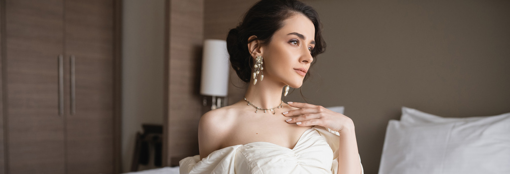 novia atractiva en vestido blanco y joyas de lujo con pendientes de perlas y collar tocando el pecho mientras está sentado en el dormitorio moderno en la habitación de hotel en el día de la boda, bandera  - Foto, Imagen