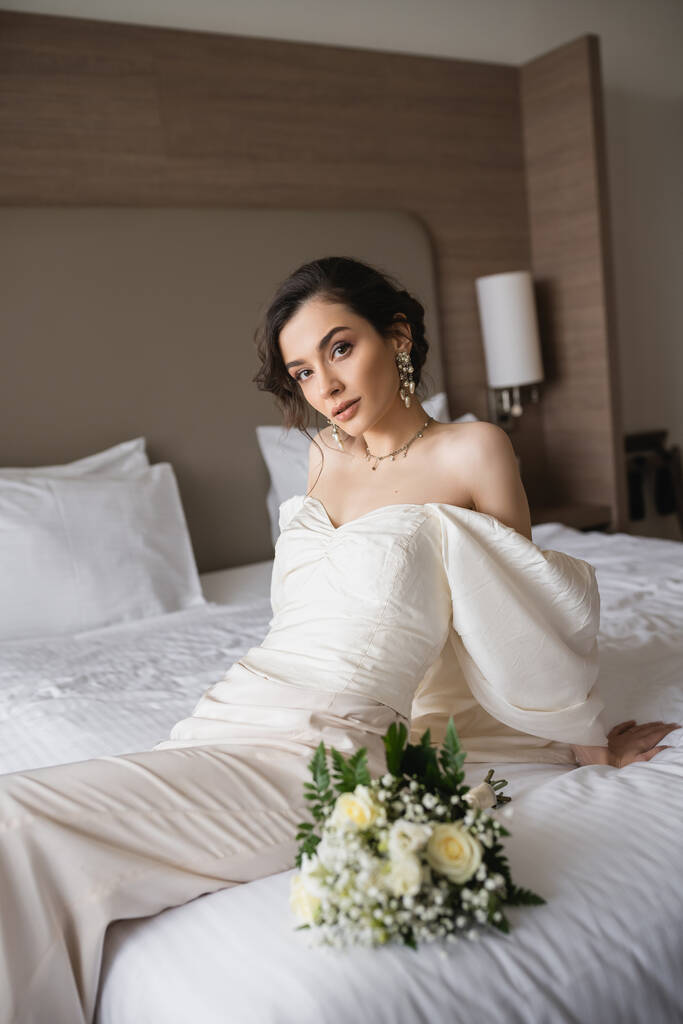 káprázatos fiatal menyasszony fehér ruhában és fényűző ékszerek ül az ágyon mellett menyasszonyi csokor virágokkal és nézi a kamera a modern hálószobában a szállodai szobában az esküvő napján  - Fotó, kép