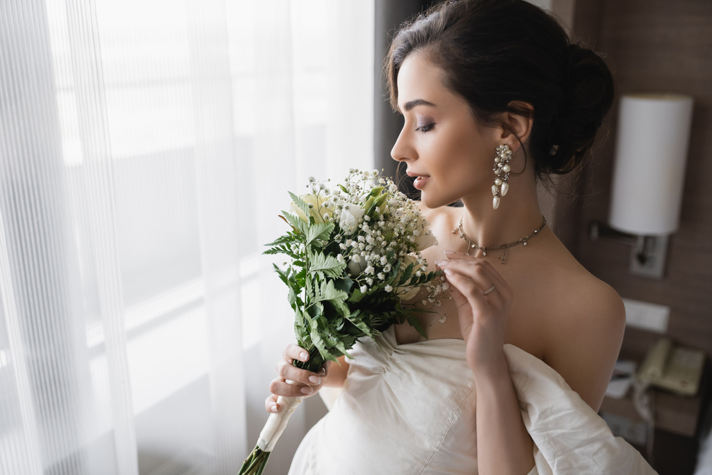 πλαϊνή άποψη της κομψής νεαρής νύφης σε λευκό φόρεμα και πολυτελή κοσμήματα με μαργαριταρένια σκουλαρίκια κρατώντας νυφικό μπουκέτο στο μοντέρνο δωμάτιο του ξενοδοχείου την ημέρα του γάμου  - Φωτογραφία, εικόνα