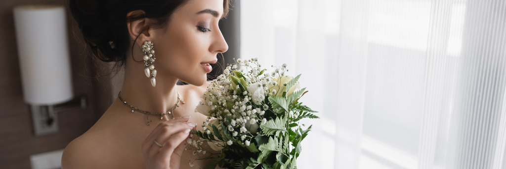 side view of elegant young bride with luxurious jewelry, in pearl earrings and necklace holding bridal bouquet in modern hotel room on wedding day, banner  - Photo, Image