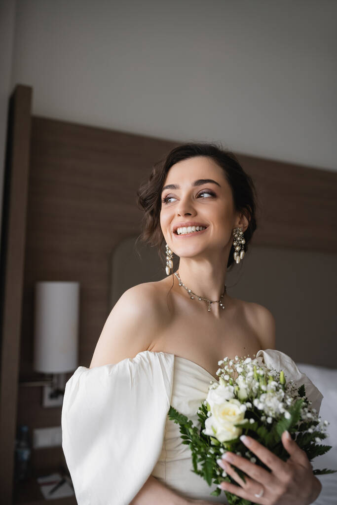 Beyaz gelinlik ve lüks takılar içinde neşeli genç bir kadın elinde çiçeklerle gelin buketi ve otel odasındaki modern yatak odasına bakıyor.  - Fotoğraf, Görsel