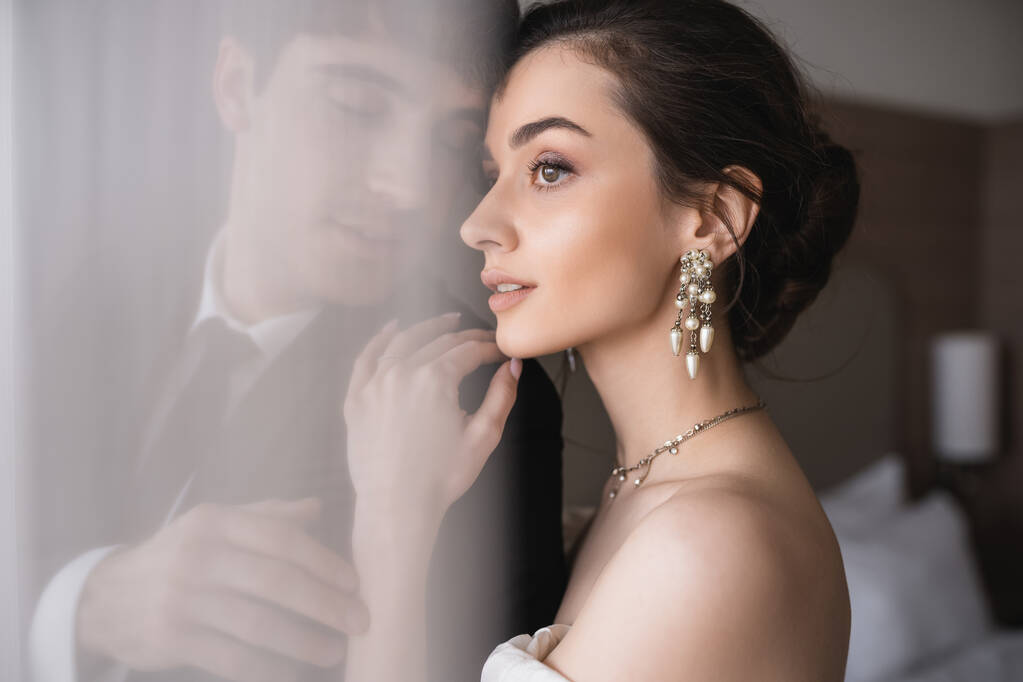 stunning bride in elegant jewelry and wedding dress hugging shoulder of groom in classic formal wear while standing together behind white tulle in modern hotel room after ceremony  - Photo, Image