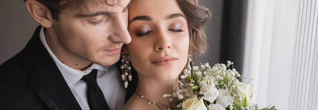 pretty bride with closed eyes, in jewelry with pearls and bridal bouquet standing next to groom in classic formal wear with tie in modern hotel room after wedding ceremony, banner  - Photo, Image