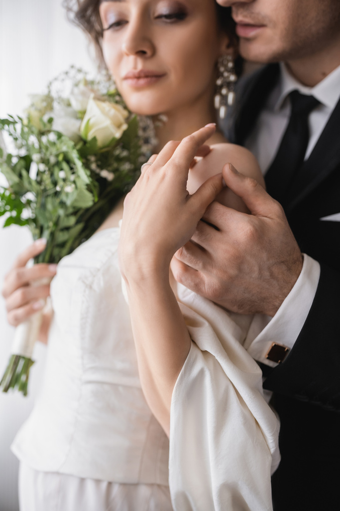 young bride in jewelry, white dress with bridal bouquet touching hands with groom in classic formal wear while standing together in modern hotel room after wedding ceremony  - Photo, Image