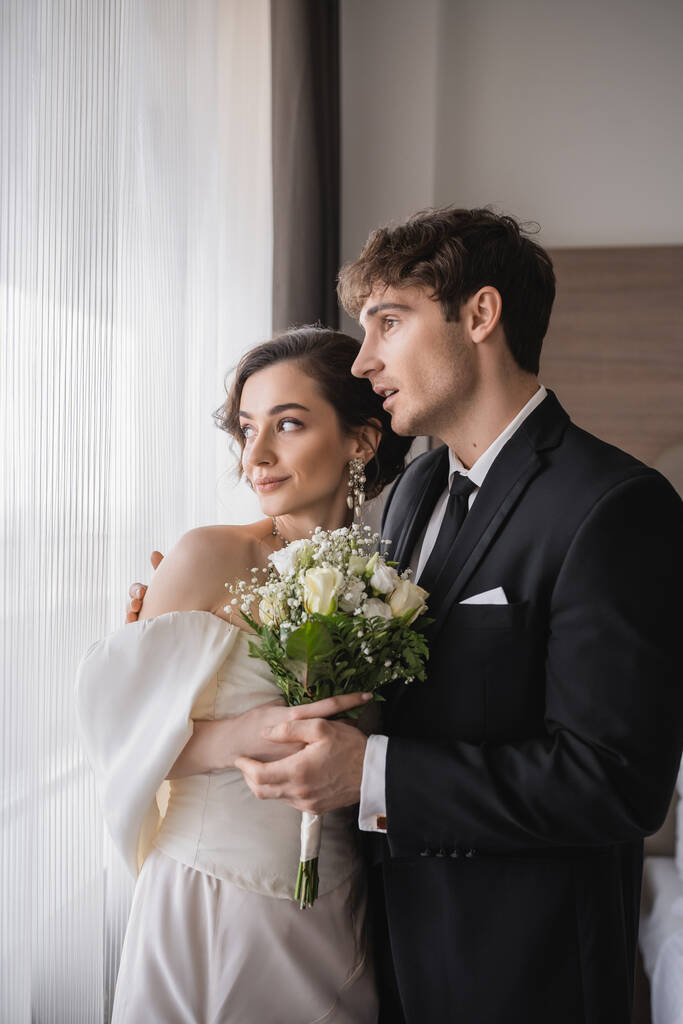užaslý ženich v klasickém formálním oblečení s otevřenými ústy vedle elegantní mladé nevěsty v špercích, bílé šaty s svatební kytice v moderním hotelovém pokoji po svatebním obřadu  - Fotografie, Obrázek