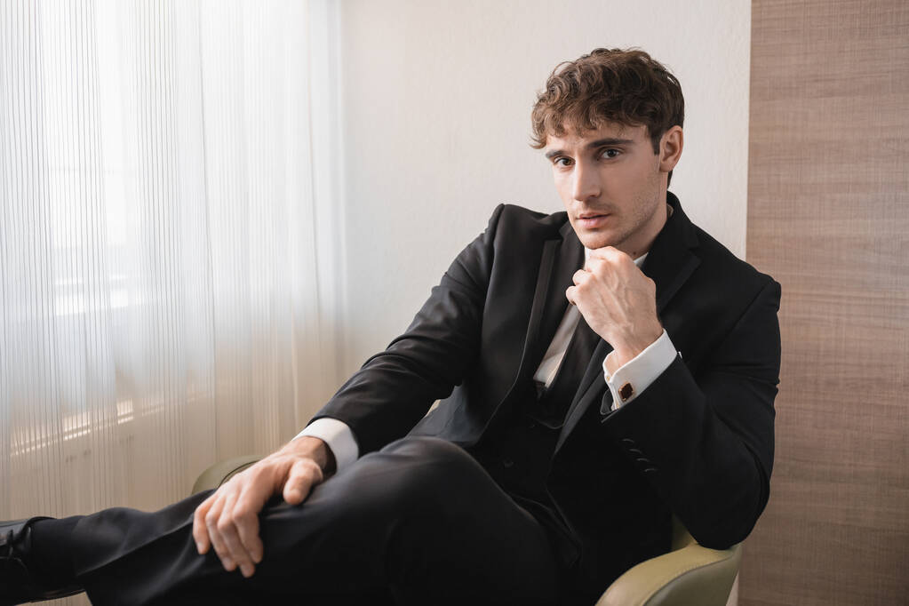 odnoszący sukcesy mężczyzna w czarnym stroju formalnym z krawatem siedzący na wygodnym fotelu i patrzący w kamerę w dniu ślubu, odpoczywający w nowoczesnym pokoju hotelowym, przystojny pan młody  - Zdjęcie, obraz