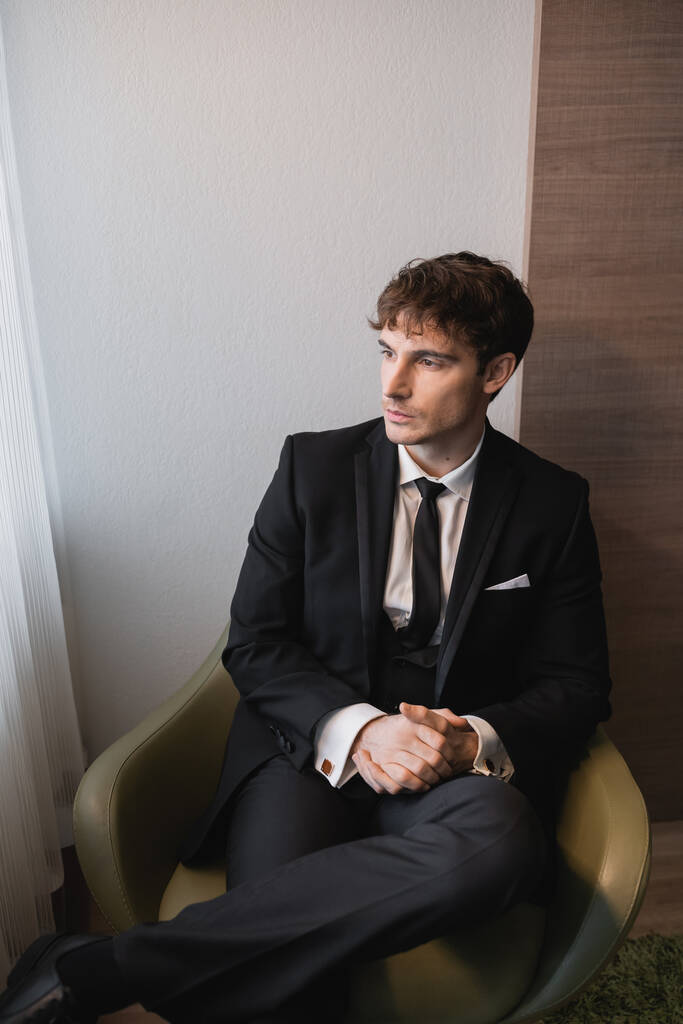 young man in black suit with tie sitting with clenched hands on comfortable armchair and looking away on wedding day, thinking in modern hotel room before marriage, good looking groom  - Photo, Image