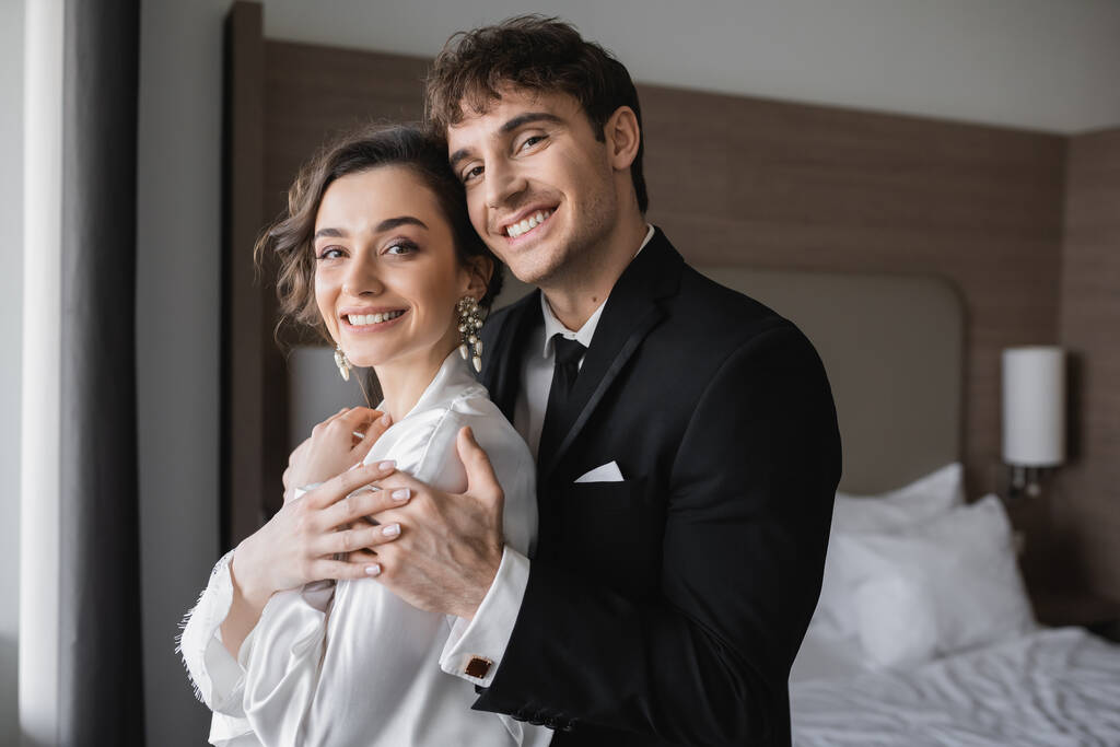 šťastný ženich v klasické formální oblečení objímající elegantní mladá nevěsta v šperky a bílé šaty při pohledu na fotoaparát společně v moderním hotelovém pokoji během jejich líbánek po svatbě  - Fotografie, Obrázek