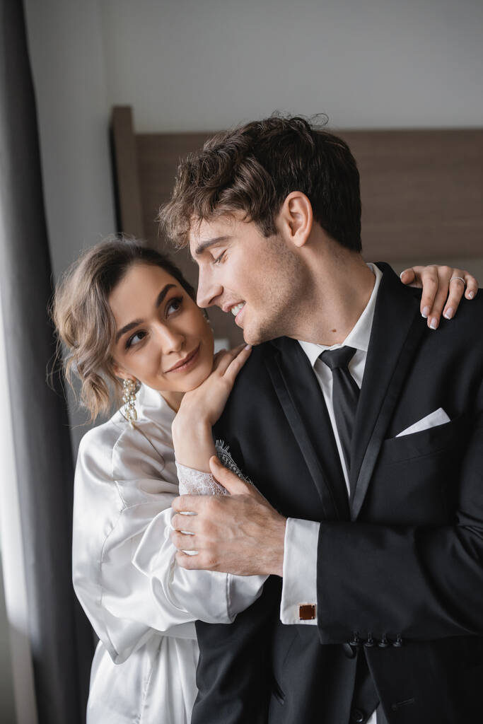 Mücevher ve beyaz ipek bornoz giymiş mutlu bir gelin klasik siyah takım elbiseli neşeli damadın omzuna yaslanırken modern otel odasında balayı sırasında yan yana duruyor, yeni evliler.  - Fotoğraf, Görsel