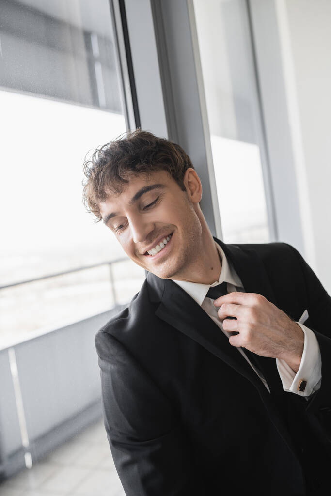 šťastný muž v klasickém společenském oblečení upravující černou kravatu při přípravě a stojící v moderním hotelovém pokoji u okna, ženich ve svatební den, zvláštní příležitosti   - Fotografie, Obrázek