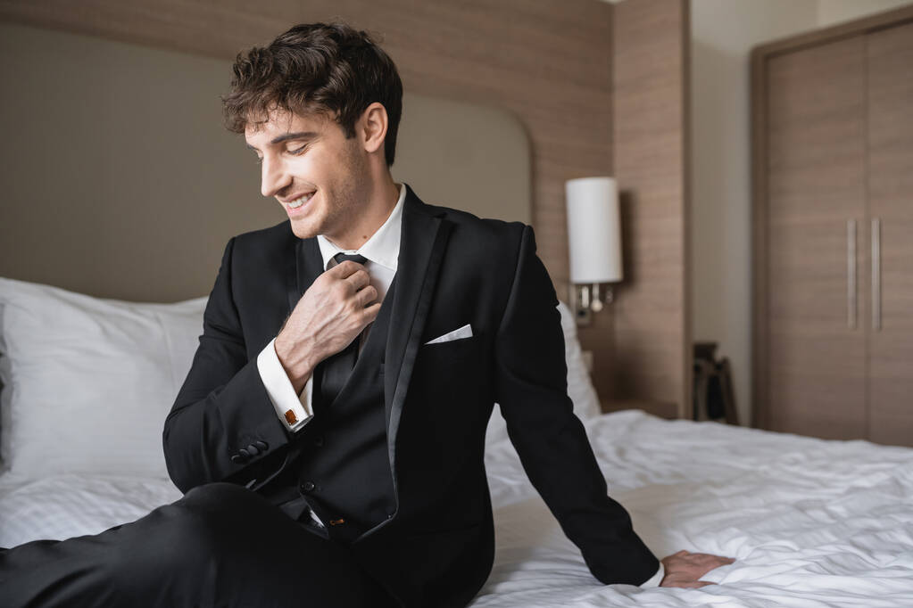 iloinen mies tyylikäs muodollinen kulumista valkoinen paita säätämällä musta solmio katsellen pois ja istuu sängyllä modernissa hotellihuoneessa, sulhanen hääpäivänä, erityinen tilaisuus   - Valokuva, kuva