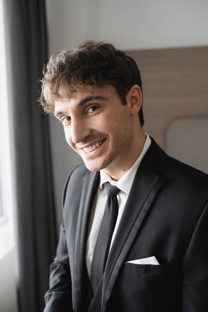 portret van een vrolijke man in stijlvolle formele kleding met zwarte stropdas en wit shirt kijkend naar camera in moderne hotelkamer, bruidegom op trouwdag, speciale gelegenheid   - Foto, afbeelding