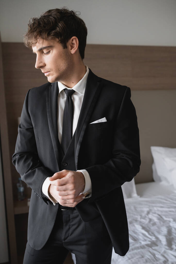 beau marié en tenue formelle avec cravate noire chic et chemise blanche debout dans la chambre d'hôtel moderne tout en ajustant les menottes, homme le jour du mariage, occasion spéciale   - Photo, image