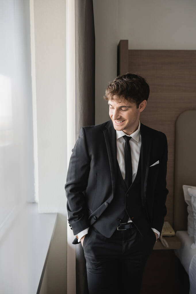 happy groom man in classy formal wear with black tie and white shirt standing with hands in pockets on pants in modern hotel room near window, man on wedding day, special occasion   - Photo, Image