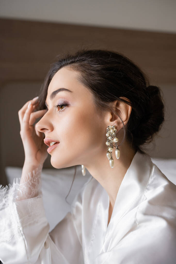 portrait of attractive woman with brunette hair in white silk robe, pearl earrings and flawless makeup preparing for her wedding in hotel room, special occasion, young bride - Photo, Image