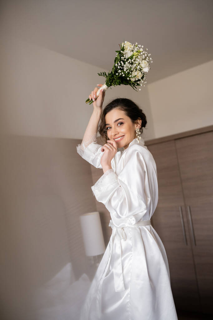 alluring young woman with brunette hair in white silk robe and pearl earrings holding bridal bouquet while preparing for her wedding in hotel room, special occasion, happy bride  - Photo, Image