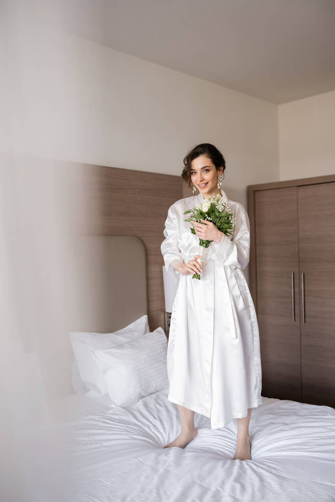 cheerful young bride with brunette hair in white silk robe and pearl earrings holding bridal bouquet while standing on bed in hotel room, special occasion, happy woman  - Photo, Image